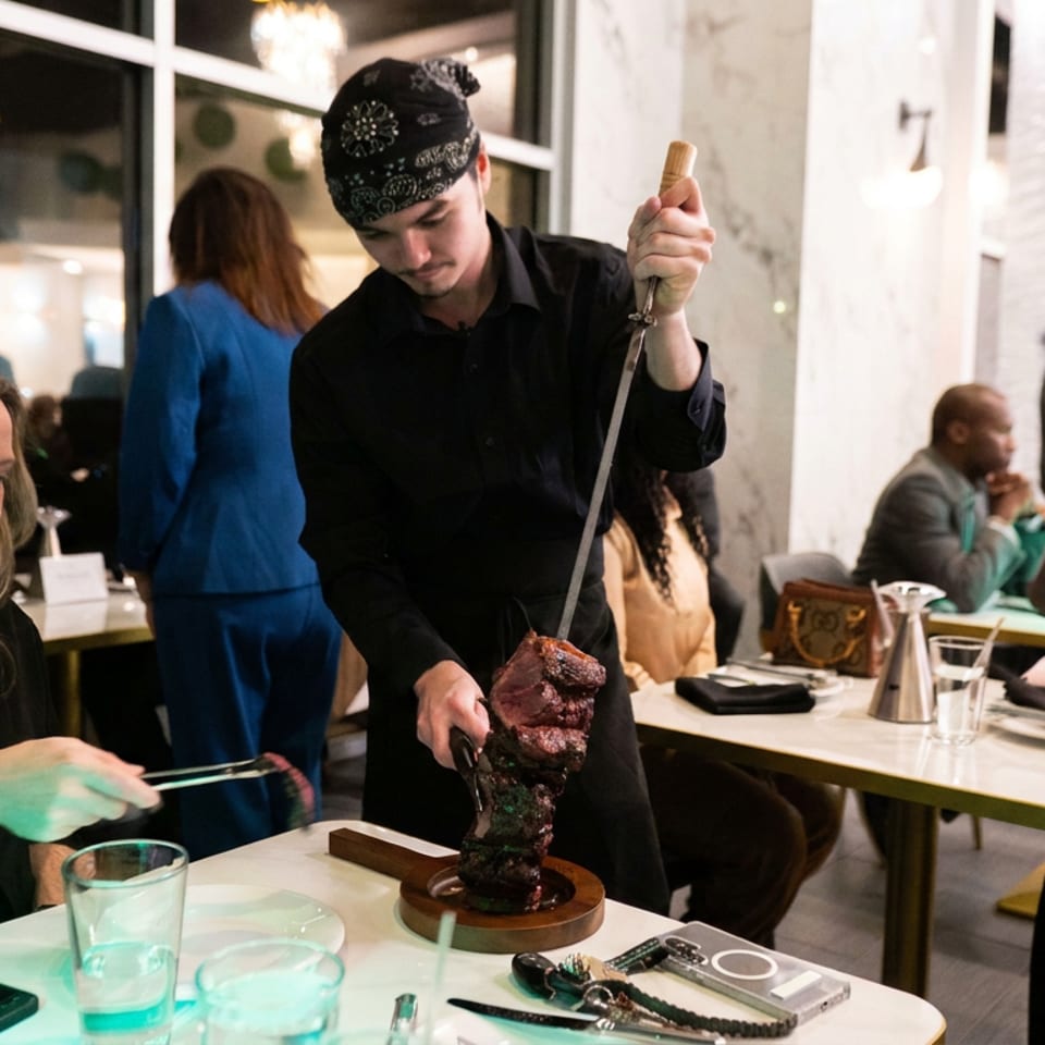 A server presents a skewer of grilled meat at a restaurant table while diners watch.