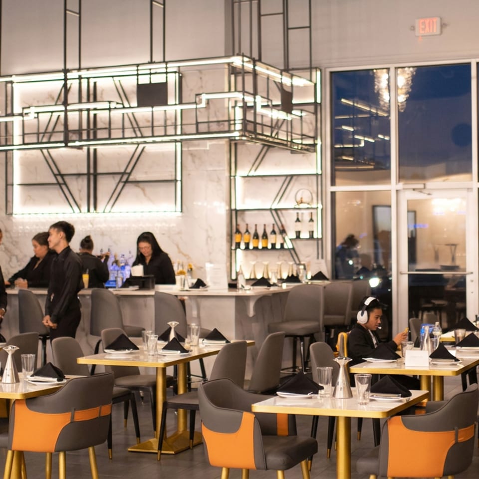 Modern upscale restaurant dining room with gold-trimmed tables, black napkins, marble bar, illuminated shelves, staff, and seated diners.