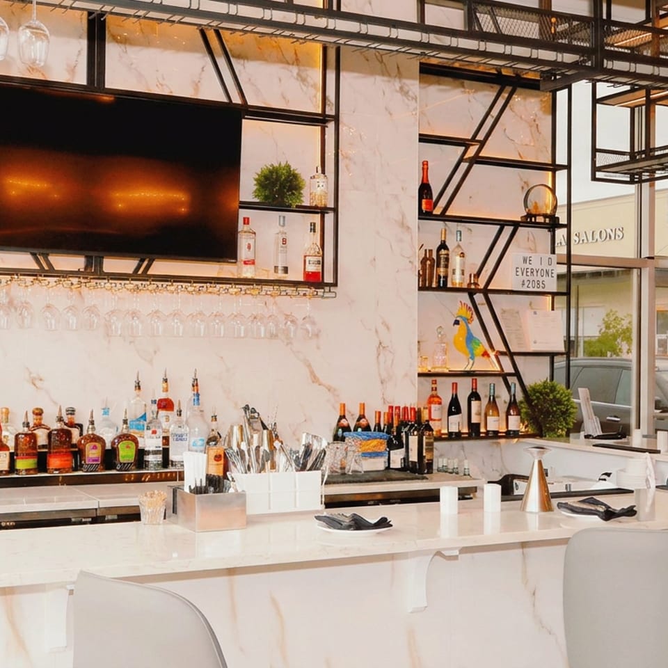 Modern marble restaurant bar with liquor bottles, hanging glassware, open shelving, television, and white counter stools.