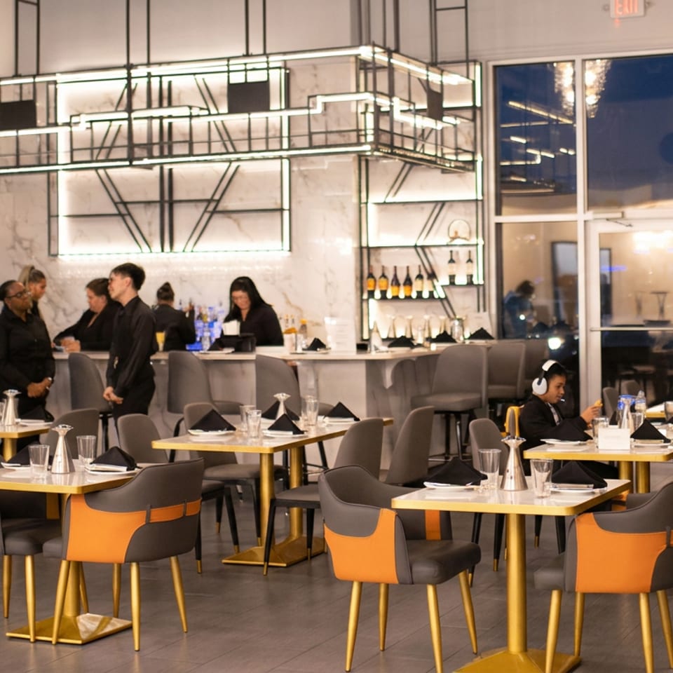 Modern restaurant dining room with orange-and-gray chairs, set tables, illuminated shelving, bar seating, and several patrons.